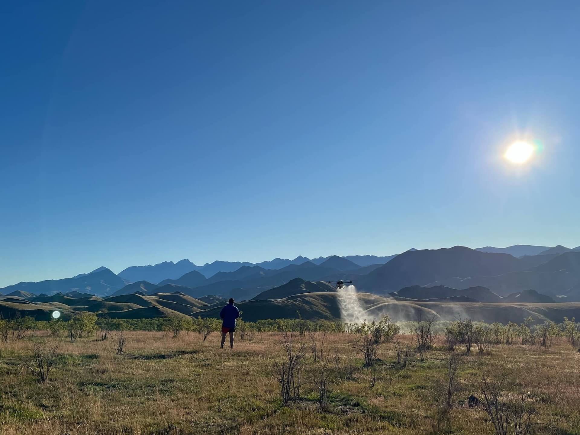 A person stands in a grassy field with distant mountains and a clear sky. A drone sprays mist nearby under the bright sun.