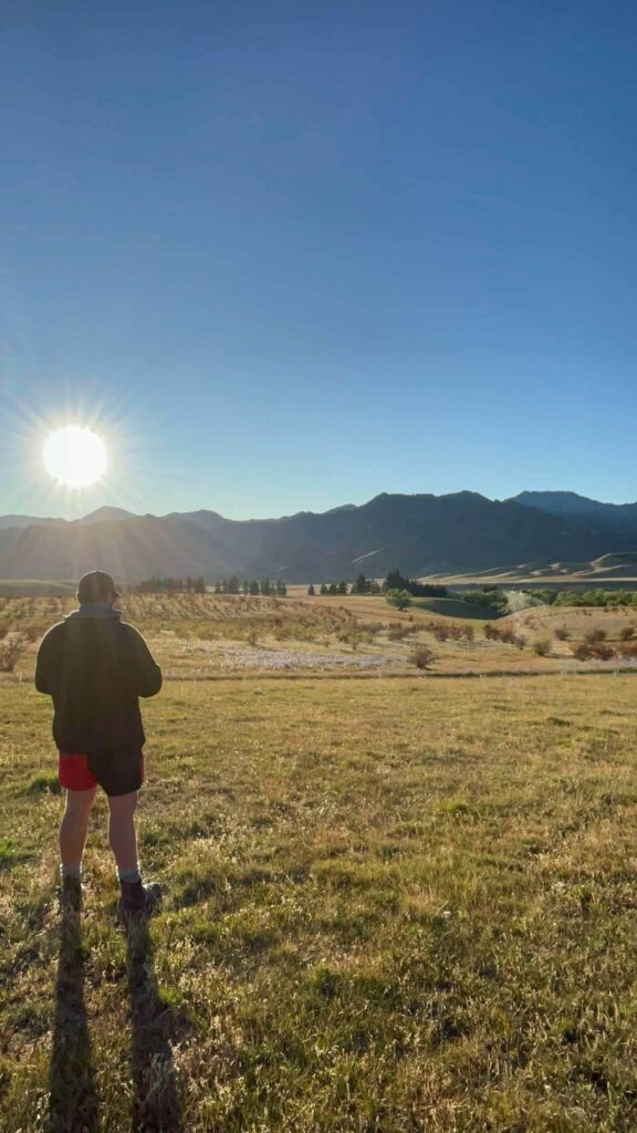Person standing controlling a drone on grassy field facing distant mountains, with the sun shining brightly in a clear blue sky.