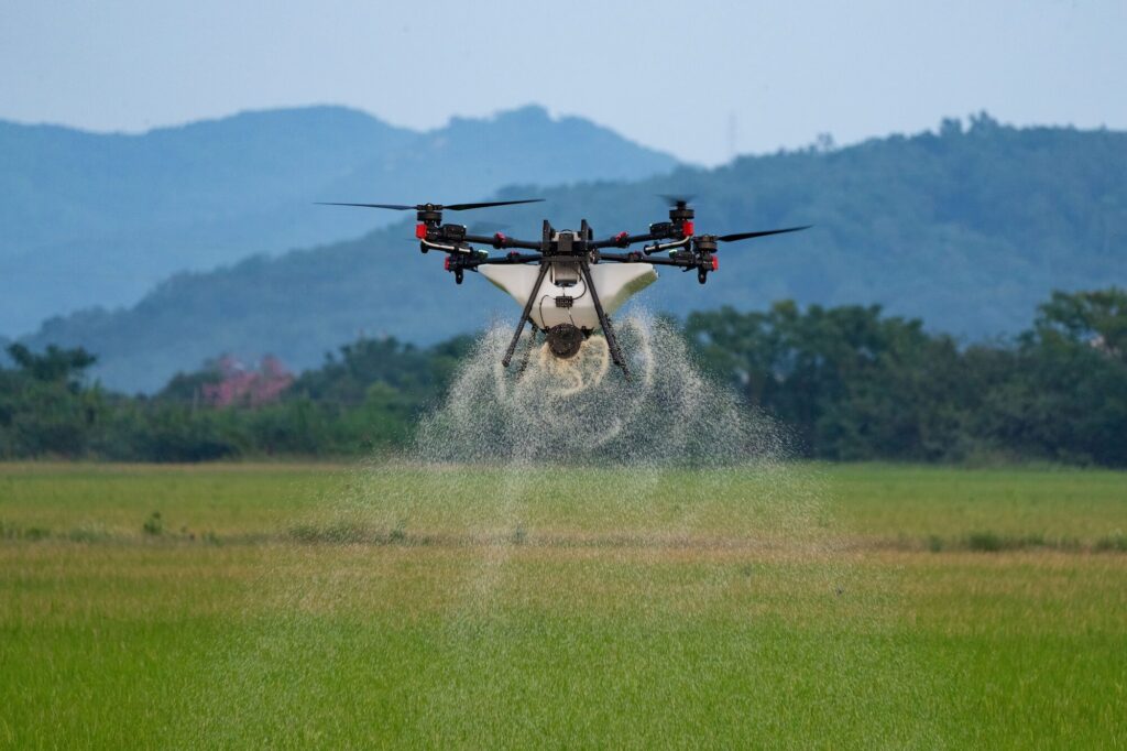 A drone sprays liquid over a green field, with hills and trees visible in the background.