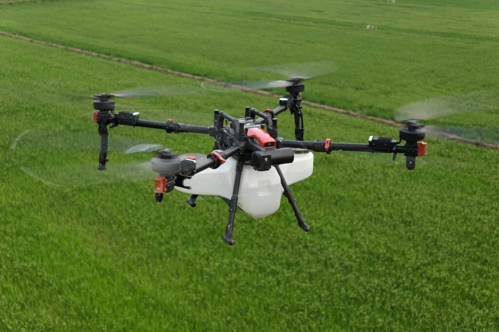 A large quadcopter drone with a white tank flies over a green agricultural field, likely used for crop spraying or monitoring.