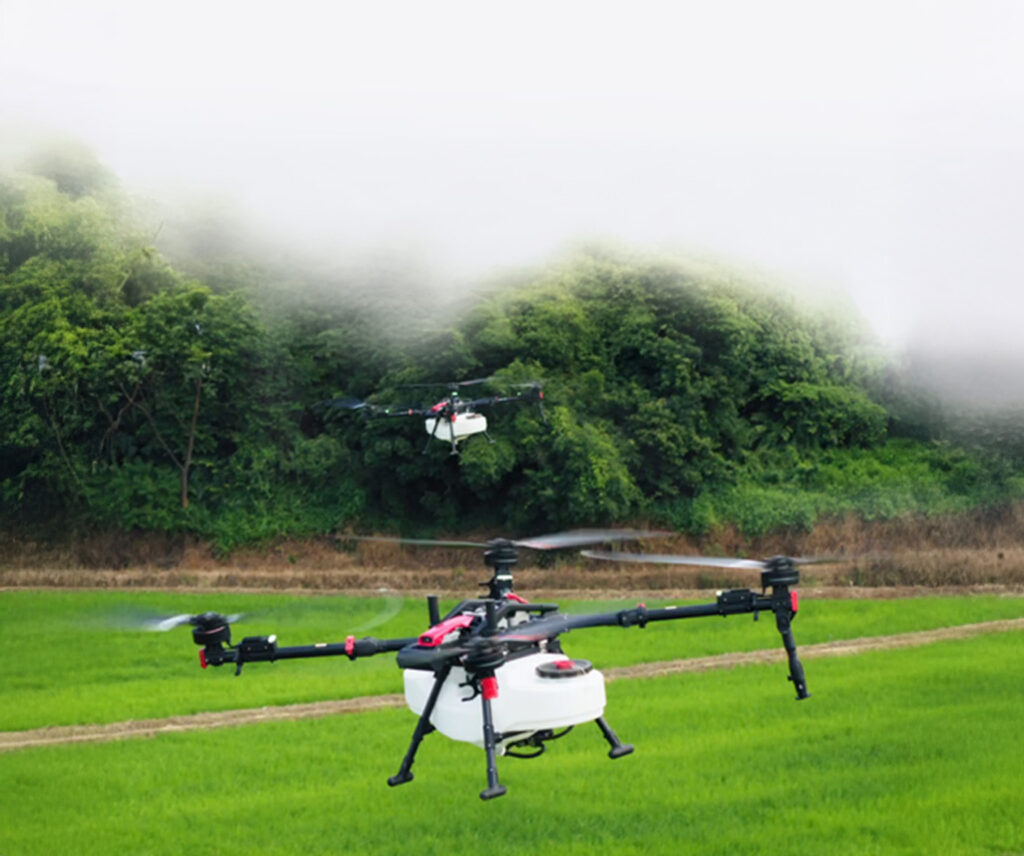Two agricultural drones hover above a green field with trees and fog in the background.